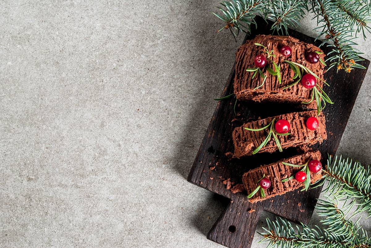 Traditional Christmas dessert, Christmas yule log cake with chocolate cream, cranberry and rosemary twigs. On stone gray background with Christmas tree branches, copy space top view